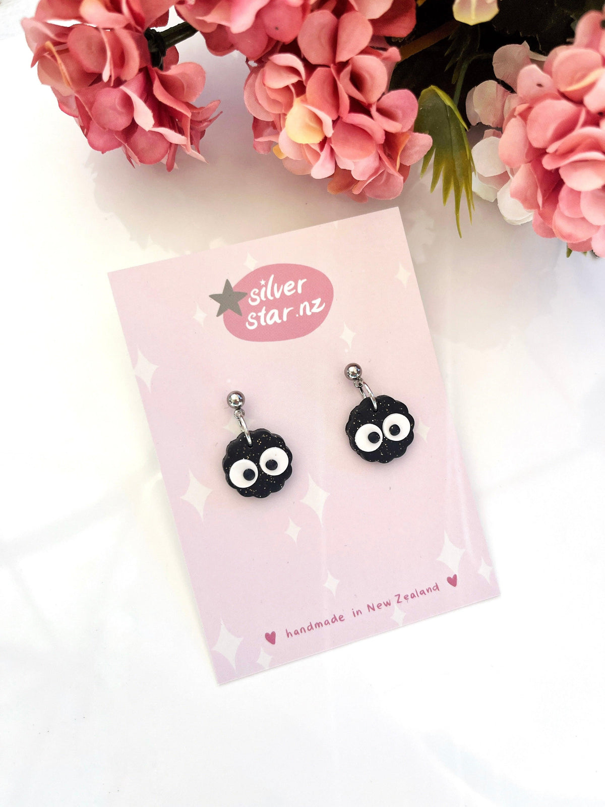 Mini Soot Star Polymer Clay Earrings by silverstar.nz feature black soot sprites with big white eyes, displayed on a pink branded card surrounded by pink flowers. The card reads "Handmade in New Zealand.