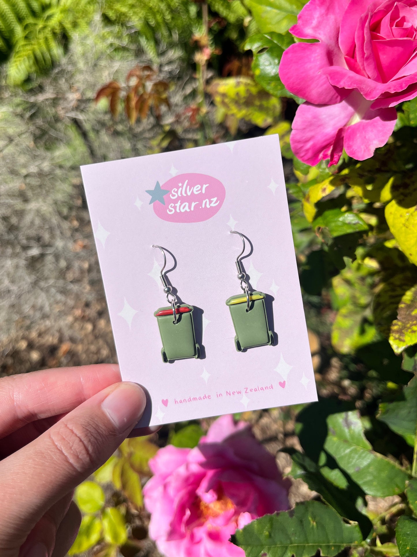 A hand holds a card from silverstar.nz displaying Rubbish Bin Polymer Clay Earrings—green bins with red and yellow lids. Pink flowers and green foliage are in the background. The card says "handmade in New Zealand.