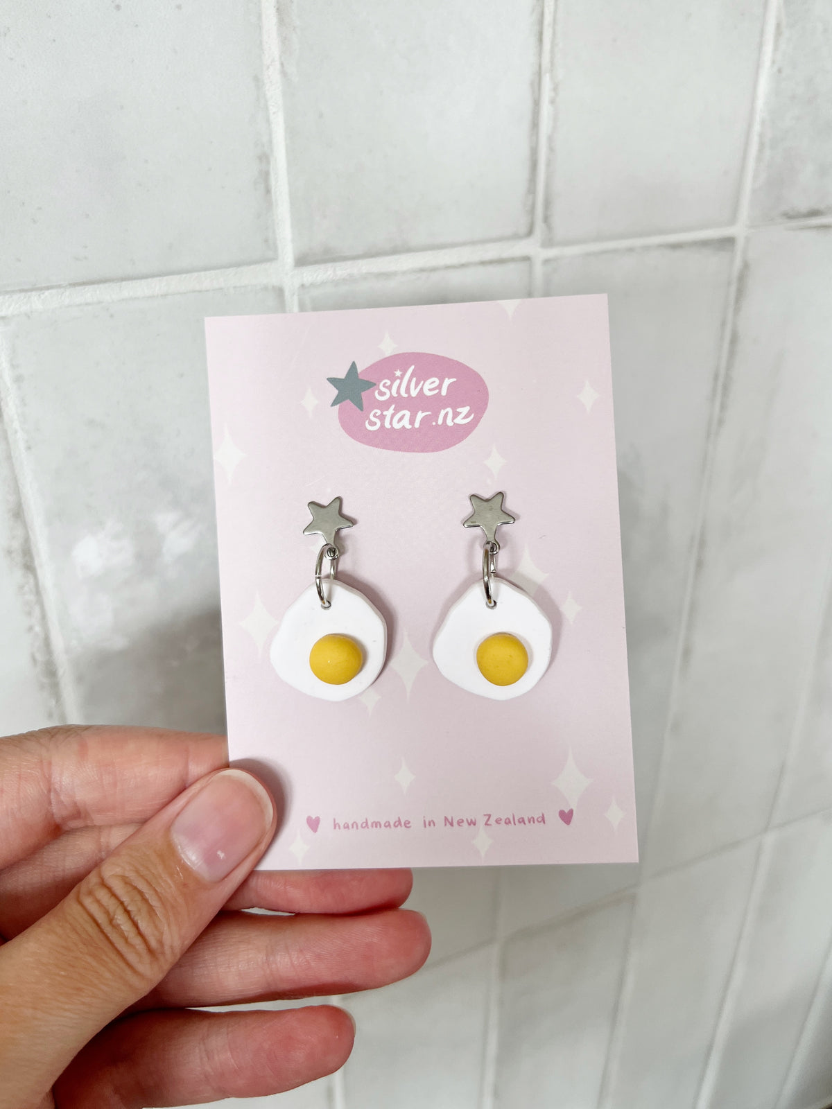 A hand holds a card from silverstar.nz displaying Egg Polymer Clay Earrings—handmade in New Zealand, featuring silver star studs and dangling white egg shapes with yellow yolks.