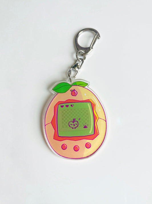 The Peach Tama Acrylic Keyring by silverstar.nz is a peach-shaped keyring with green leaves, a digital pet screen displaying a pixel creature and three hearts, plus three pink buttons—perfect as a cute accessory.