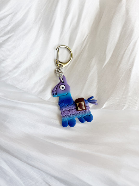 The Llama Polymer Clay Keyring by silverstar.nz, shaped like a vibrant llama piñata in purple, blue, and teal, rests on white textured fabric—a charming pick for unique gifts.