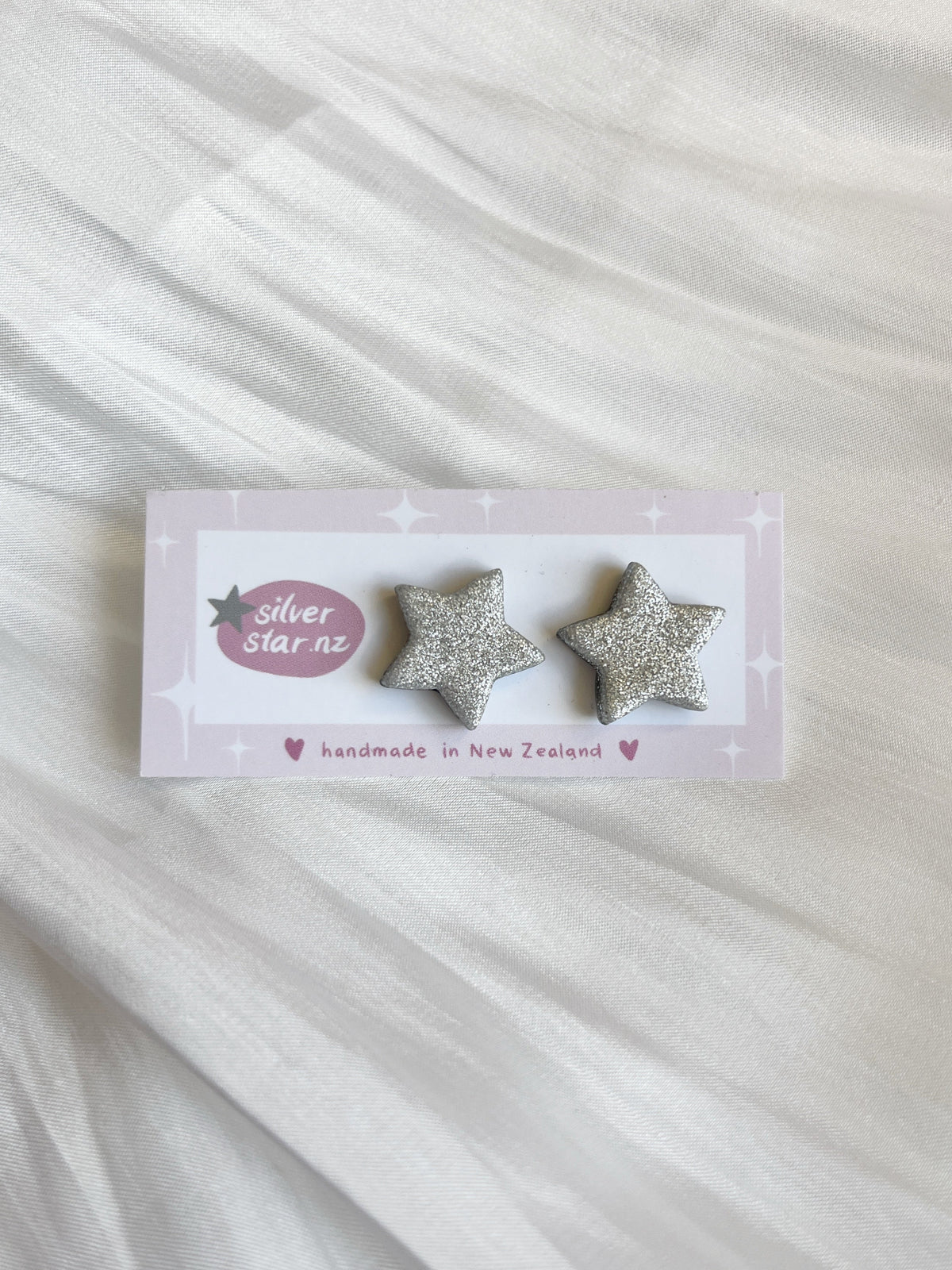 The Silver Star Polymer Clay Stud Earrings feature a unique glittery silver star design, displayed on a light pink card labeled silverstar.nz and handmade in New Zealand, resting elegantly on soft white fabric.