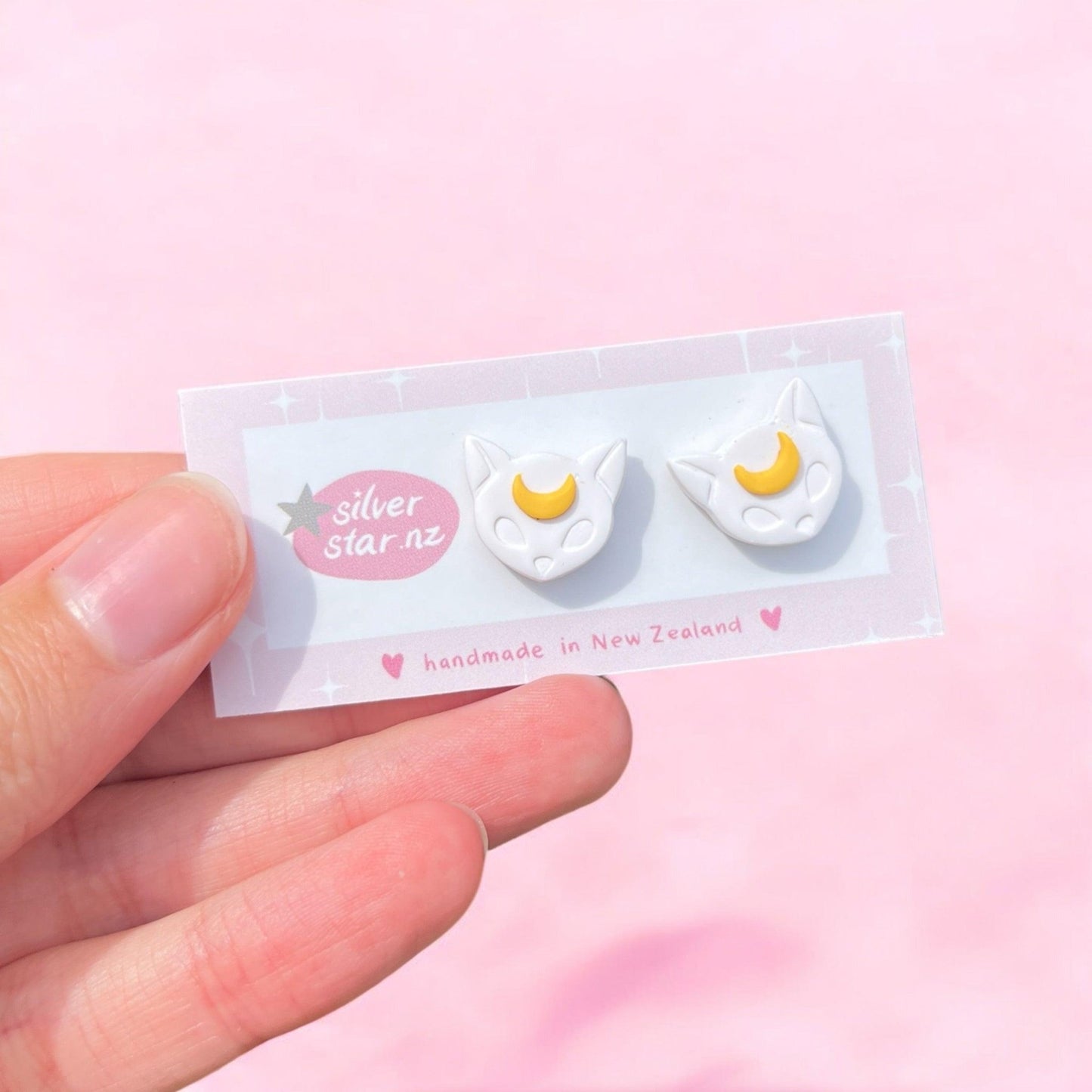 A hand holds a card showing Moon Cat Polymer Clay Stud Earrings—white cat-shaped studs with yellow crescent moons—from silverstar.nz, labeled "handmade in New Zealand," against a soft pink background.