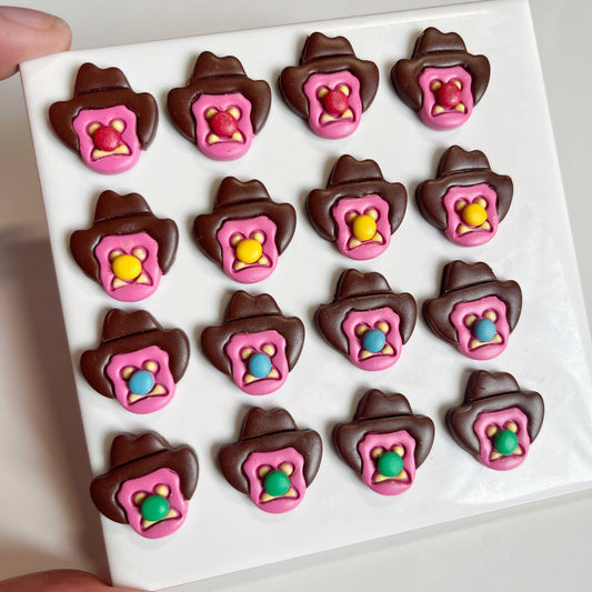 A white tray displays 16 Bubble 'O Will Polymer Clay Earrings by silverstar.nz—brown cowboy hat shapes with pink trim and a colorful candy center (yellow, blue, green, or pink), resembling playful handmade sweets.