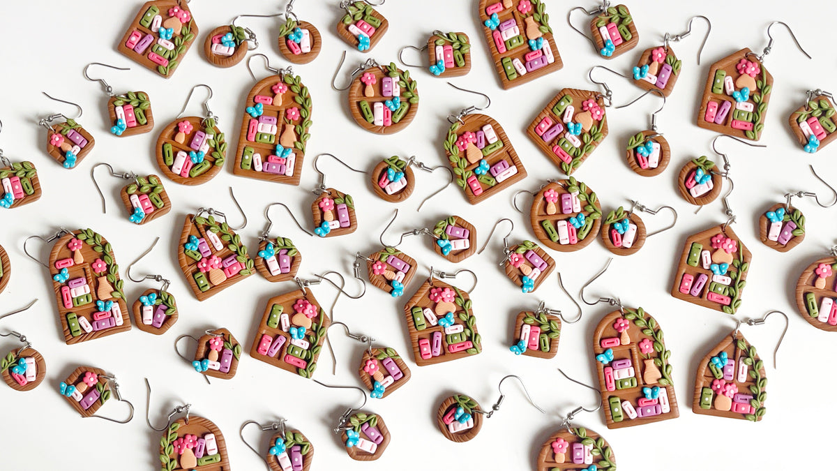 A collection of colorful, handmade clay earrings and pendants shaped like houses with pink, blue, and green details, displayed on a white background.