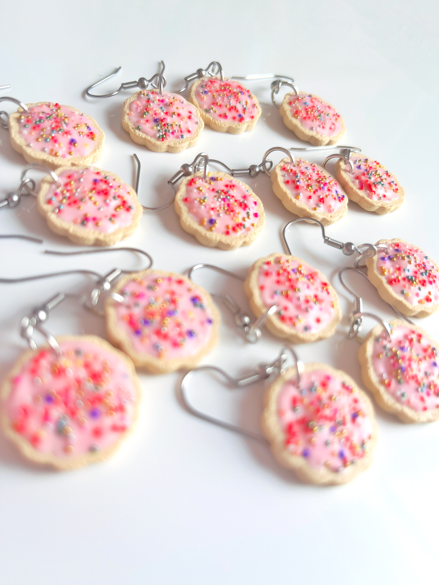 Several pairs of earrings designed to look like pink frosted cookies with colorful sprinkles are scattered on a white surface.