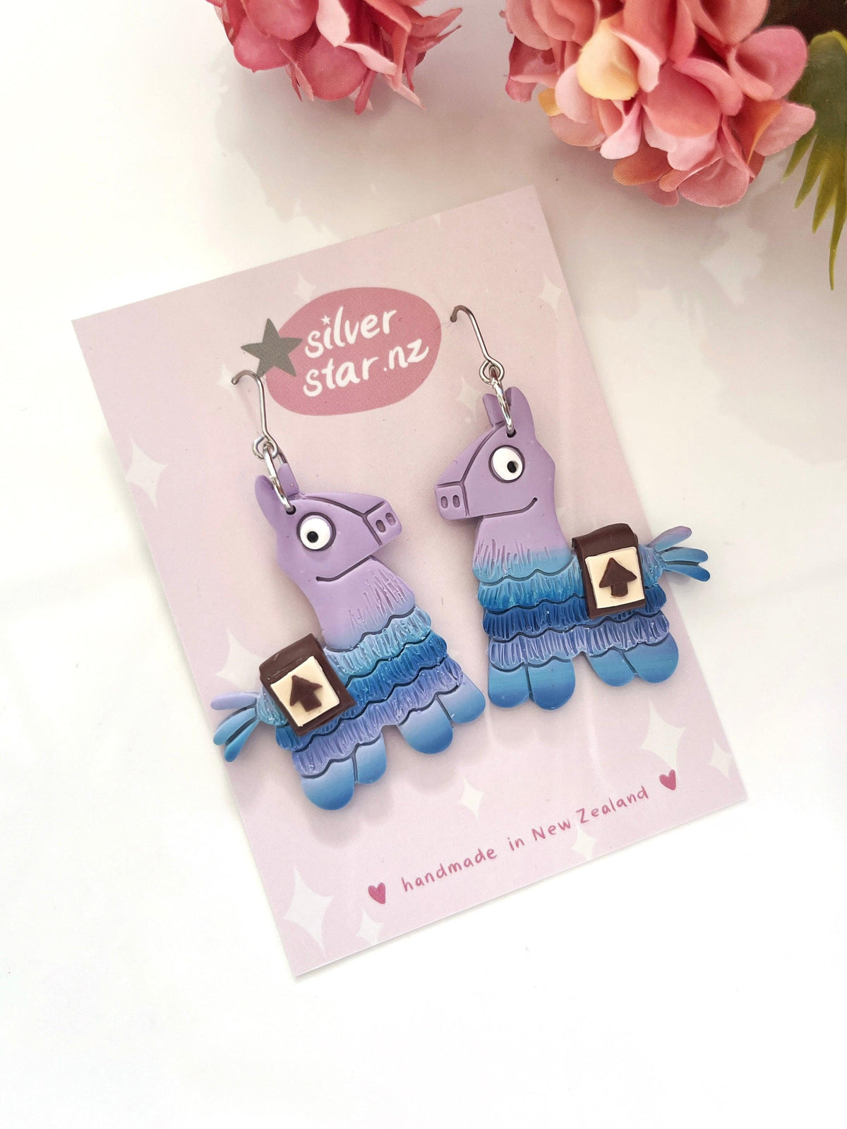 Llama Polymer Clay Earrings by silverstar.nz—handmade in purple and blue cartoon llama shapes, displayed on a pink silverstar.nz card with pink flowers. Unique New Zealand earrings to brighten any collection.