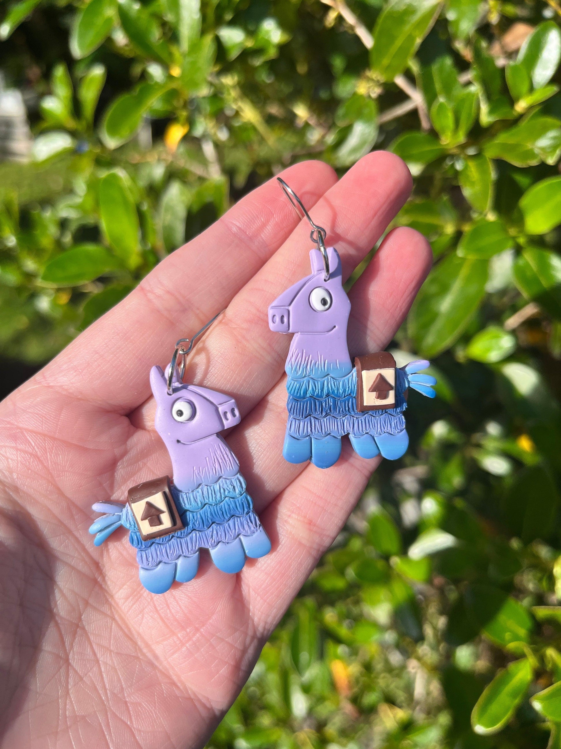 A hand displays two Llama Polymer Clay Earrings by silverstar.nz in purple and blue, each with a small box detail. Bright green leaves are visible in the sunny background.
