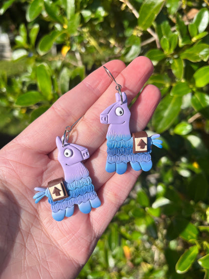 A hand displays two Llama Polymer Clay Earrings by silverstar.nz in purple and blue, each with a small box detail. Bright green leaves are visible in the sunny background.