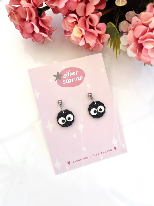 Mini Soot Star Polymer Clay Earrings by silverstar.nz feature black soot sprites with big white eyes, displayed on a pink branded card surrounded by pink flowers. The card reads "Handmade in New Zealand.
