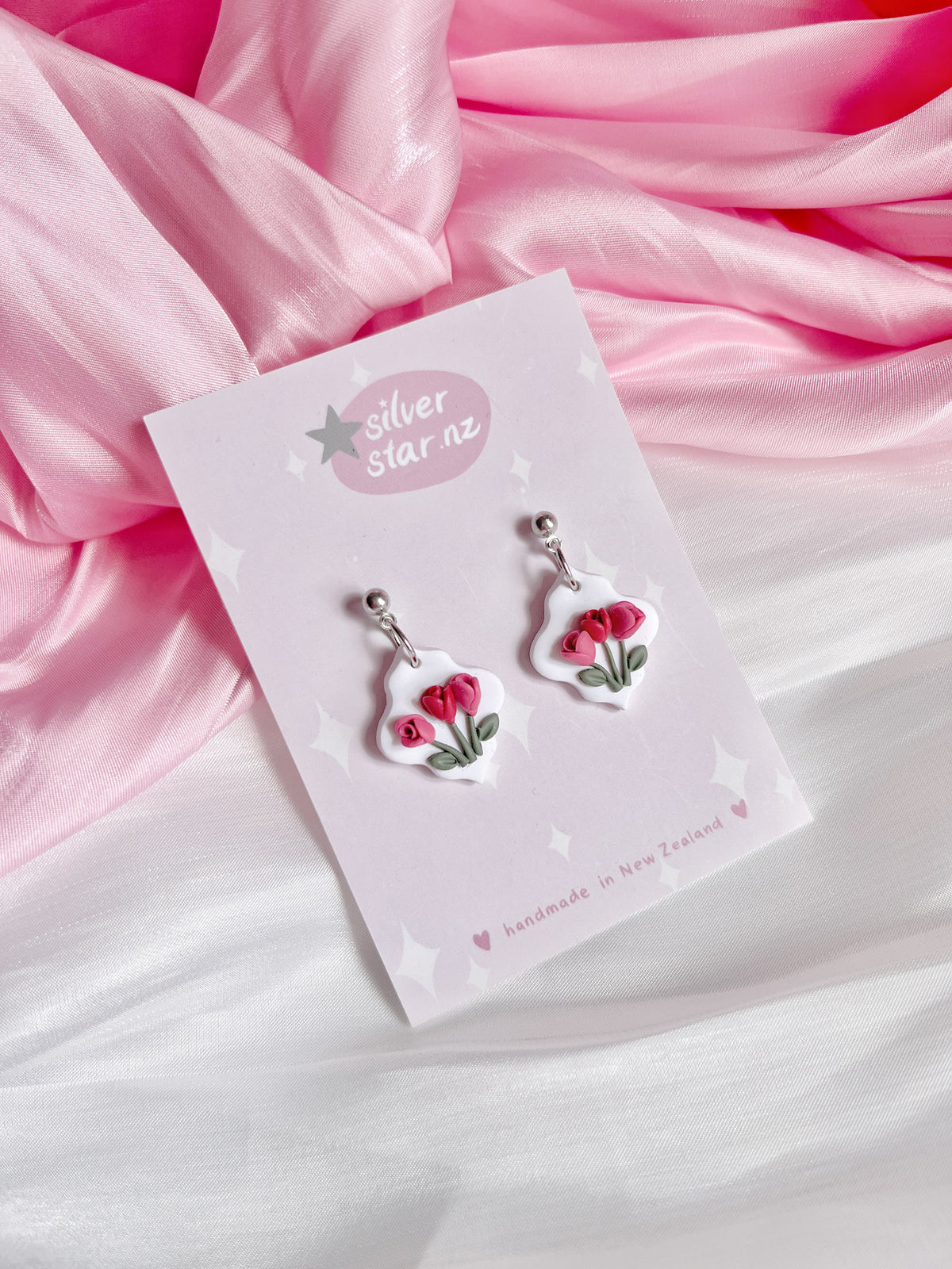 The Tulip Floral Polymer Clay Earrings, featuring pink flower pendants, are elegantly displayed on a card labeled silverstar.nz, resting on draped pink and white fabric. These silver earrings are handmade in New Zealand.