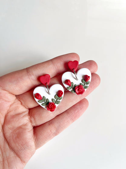 A hand showcases silverstar.nzs Heart Rose Floral Polymer Clay Earrings, featuring funky heart-shaped designs with red roses and green leaves on a white background, topped with vibrant red heart studs.