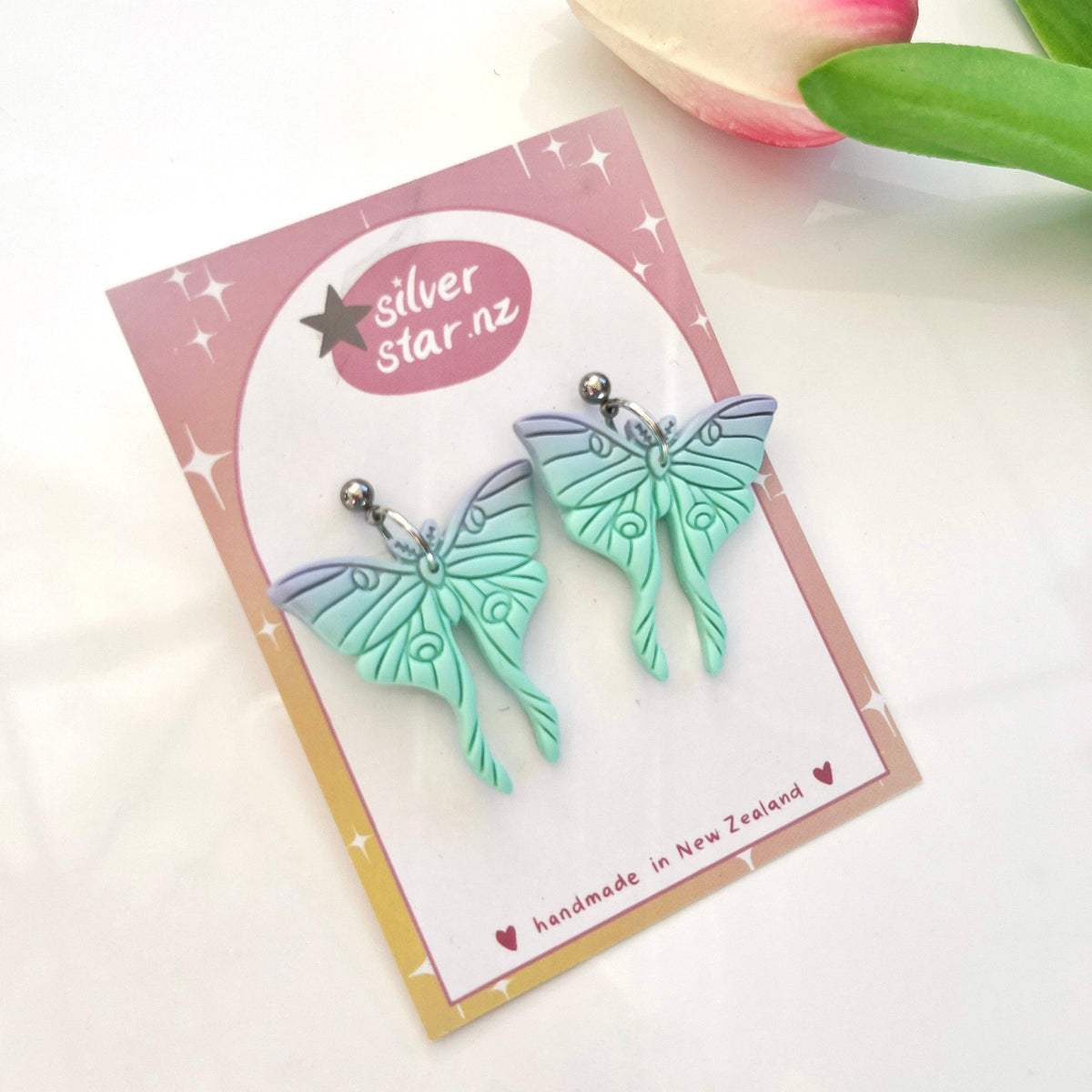 A pair of mint green Moth Polymer Clay Earrings with intricate designs is displayed on a card. The card reads "silver star.nz," indicating they are handmade in New Zealand. Crafted from polymer clay and featuring stainless steel hooks, a pink tulip is partially visible in the background.