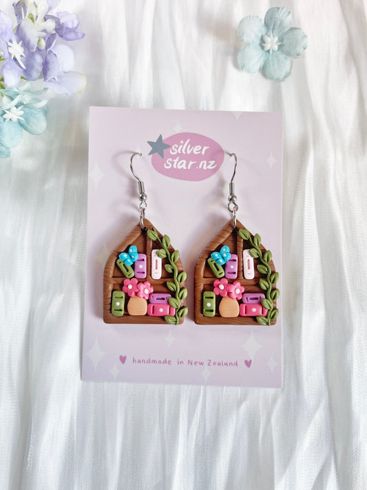 Pointed Arch Bookshelf Polymer Clay Earrings by silverstar.nz feature brown house shapes with pink flowers, greenery, and colorful rectangles on stainless steel hooks. Displayed on a silverstar.nz card against a white floral background.