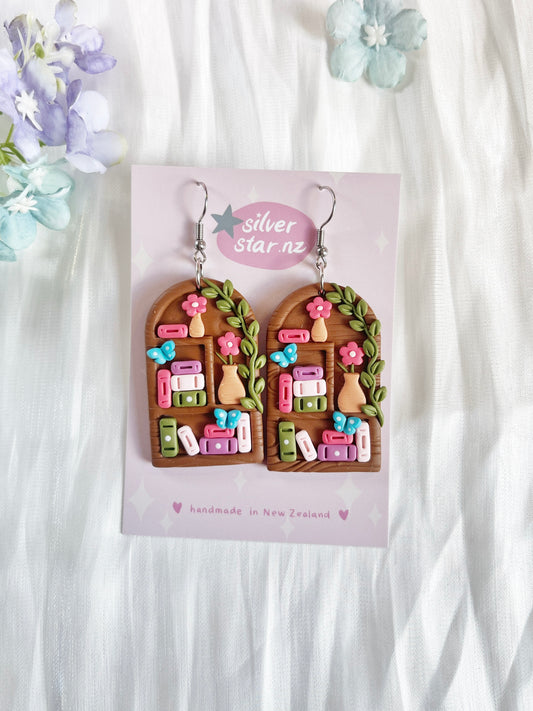 Tall Arch Bookshelf Polymer Clay Earrings by silverstar.nz feature colorful books, potted plants, and pink flowers on arched shapes with stainless steel hooks. Presented on a branded card with a floral background.