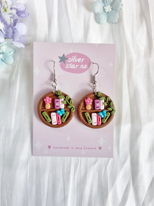 Round Bookshelf Polymer Clay Earrings by silverstar.nz—handmade, round earrings featuring colorful miniature books, flowers, and leaves. Displayed on a branded card with pastel stars and hearts for a unique look.