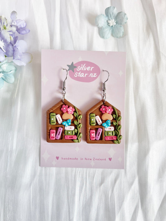 Cottage Bookshelf Polymer Clay Earrings by silverstar.nz feature a house-shaped design with colorful mini books, plants, and flowers. Displayed on a handmade in New Zealand card, pastel flowers rest on white fabric nearby.