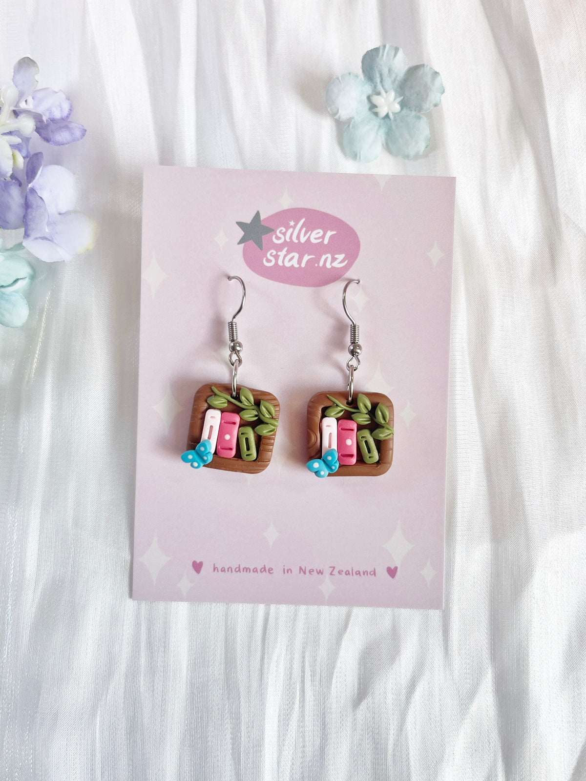 Explore silverstar.nzs Mini Square Bookshelf Polymer Clay Earrings, featuring funky miniature book designs with tiny leaves and blue flowers. With stainless steel hooks and presented on a floral pink card, these handcrafted earrings blend style and charm.
