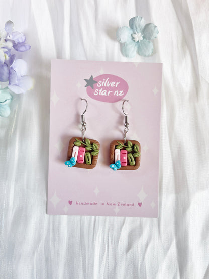 Explore silverstar.nzs Mini Square Bookshelf Polymer Clay Earrings, featuring funky miniature book designs with tiny leaves and blue flowers. With stainless steel hooks and presented on a floral pink card, these handcrafted earrings blend style and charm.