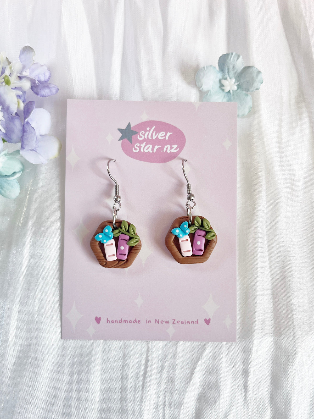 Mini Hexagon Bookshelf Polymer Clay Earrings feature baskets with handmade clay, pink and white objects, brown handles, and blue flowers. The card reads silverstar.nz with handmade in New Zealand. These unique earrings stand out against light fabric and pastel flowers.