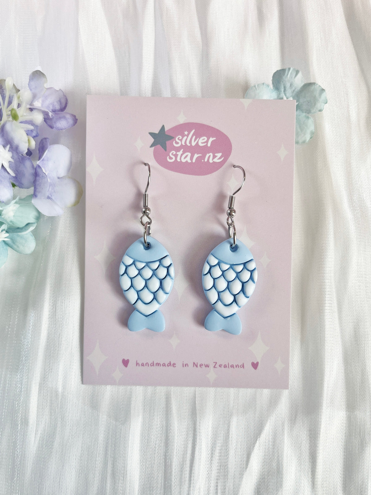 The Fishy Polymer Clay Earrings, shaped like blue fish with white scales and hanging from hooks, are displayed on a card labeled silverstar.nz and handmade in New Zealand, resting on white and light blue fabric with purple and blue flowers.