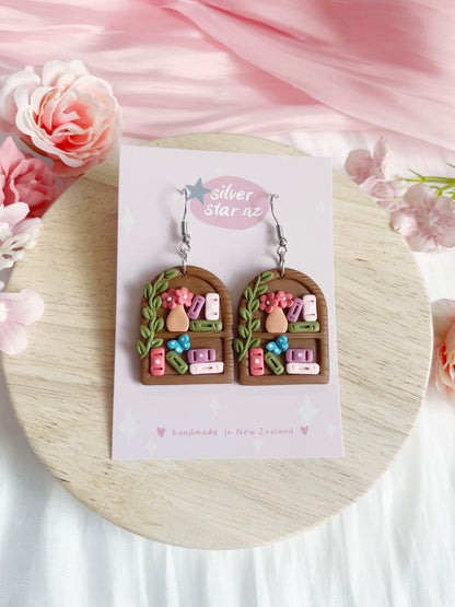 The Round Arch Bookshelf Polymer Clay Earrings by silverstar.nz feature brown arched bookshelf shapes with colorful mini books, plant vine, flowers, vase details, stainless steel hooks, and come on a branded pink-floral backing card.