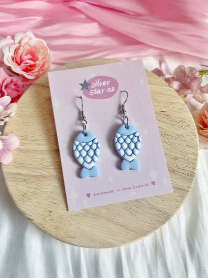 The Fishy Polymer Clay Earrings, from silverstar.nz, feature unique blue fish designs with scale patterns. They rest on a branded card on wood, framed by pink fabric and flowers, showcasing their handmade craftsmanship in New Zealand.