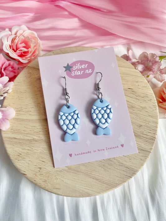 The Fishy Polymer Clay Earrings, from silverstar.nz, feature unique blue fish designs with scale patterns. They rest on a branded card on wood, framed by pink fabric and flowers, showcasing their handmade craftsmanship in New Zealand.