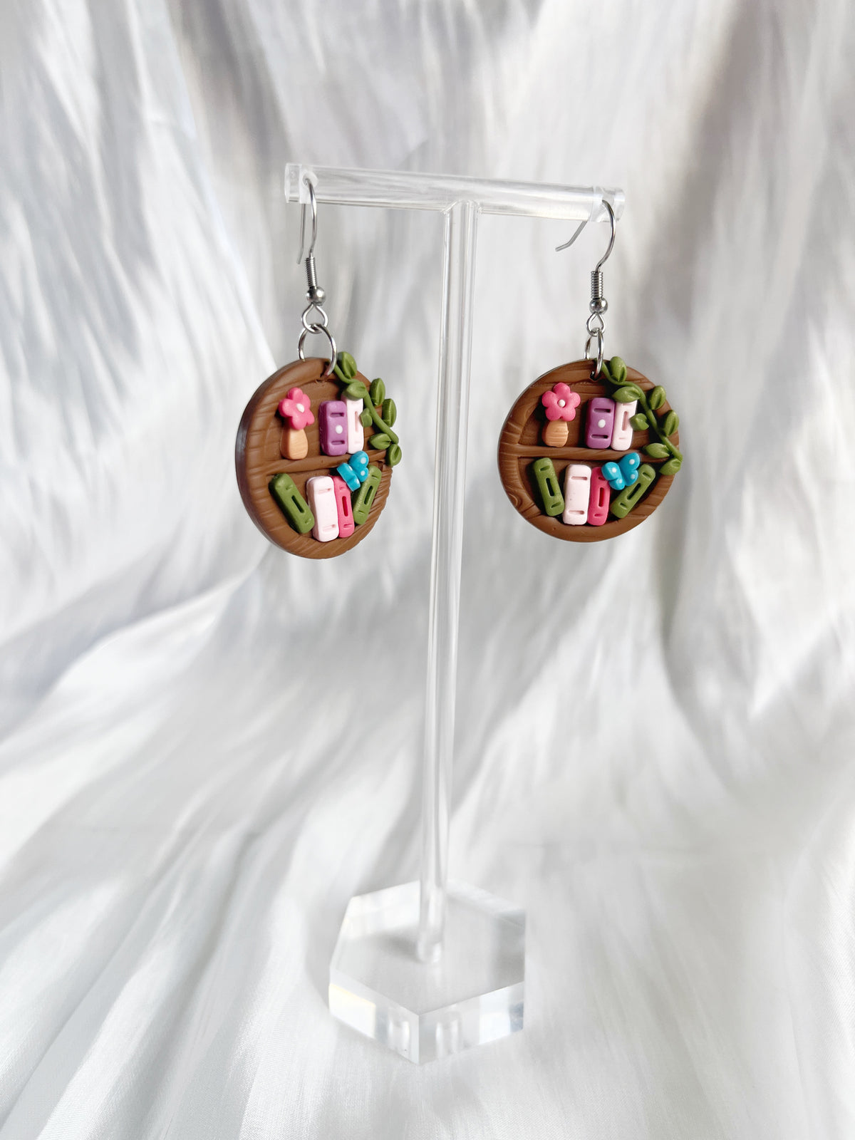 Round Bookshelf Polymer Clay Earrings by silverstar.nz are handmade, featuring miniature books, flowers, and leaves on a wooden-look base. Displayed on a clear stand against a soft white textured fabric.