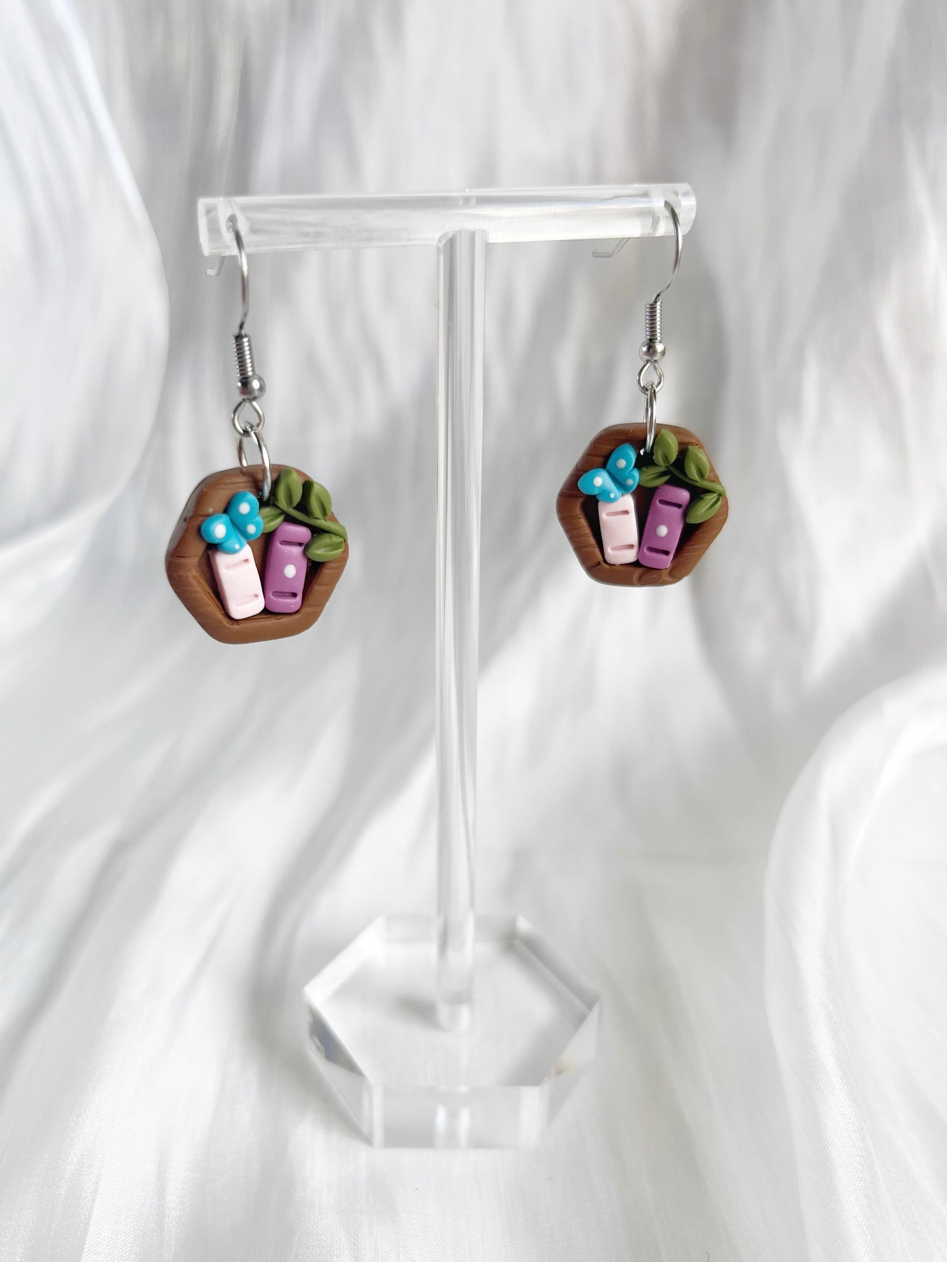 The Mini Hexagon Bookshelf Polymer Clay Earrings by silverstar.nz are showcased on a clear stand with a soft white background. Featuring tiny blue and purple book charms with green leaf accents, these whimsical earrings use stainless steel hooks for lasting wear.