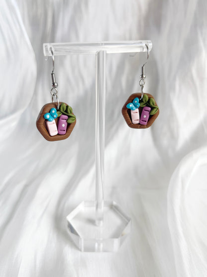 The Mini Hexagon Bookshelf Polymer Clay Earrings by silverstar.nz are showcased on a clear stand with a soft white background. Featuring tiny blue and purple book charms with green leaf accents, these whimsical earrings use stainless steel hooks for lasting wear.
