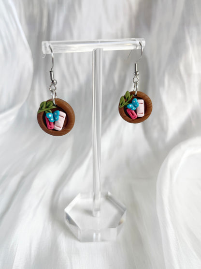 The Mini Round Bookshelf Polymer Clay Earrings by silverstar.nz feature a unique wooden plate design with miniature sushi rolls, including pink fish and green leaves, displayed on a transparent stand against a white background.