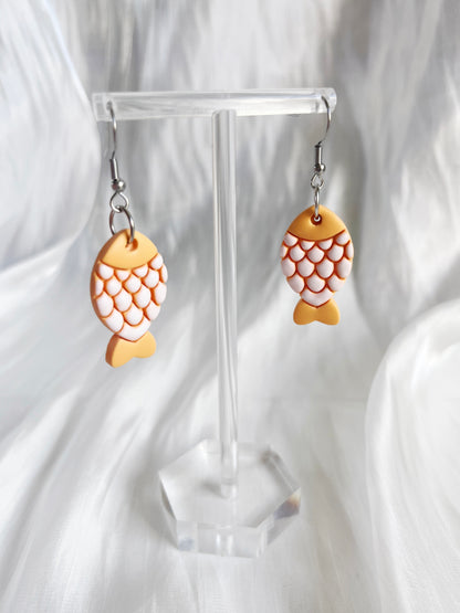 Fishy Polymer Clay Earrings by silverstar.nz feature orange and white scales, elegantly displayed on a transparent stand against a silky white background, highlighting their unique design.