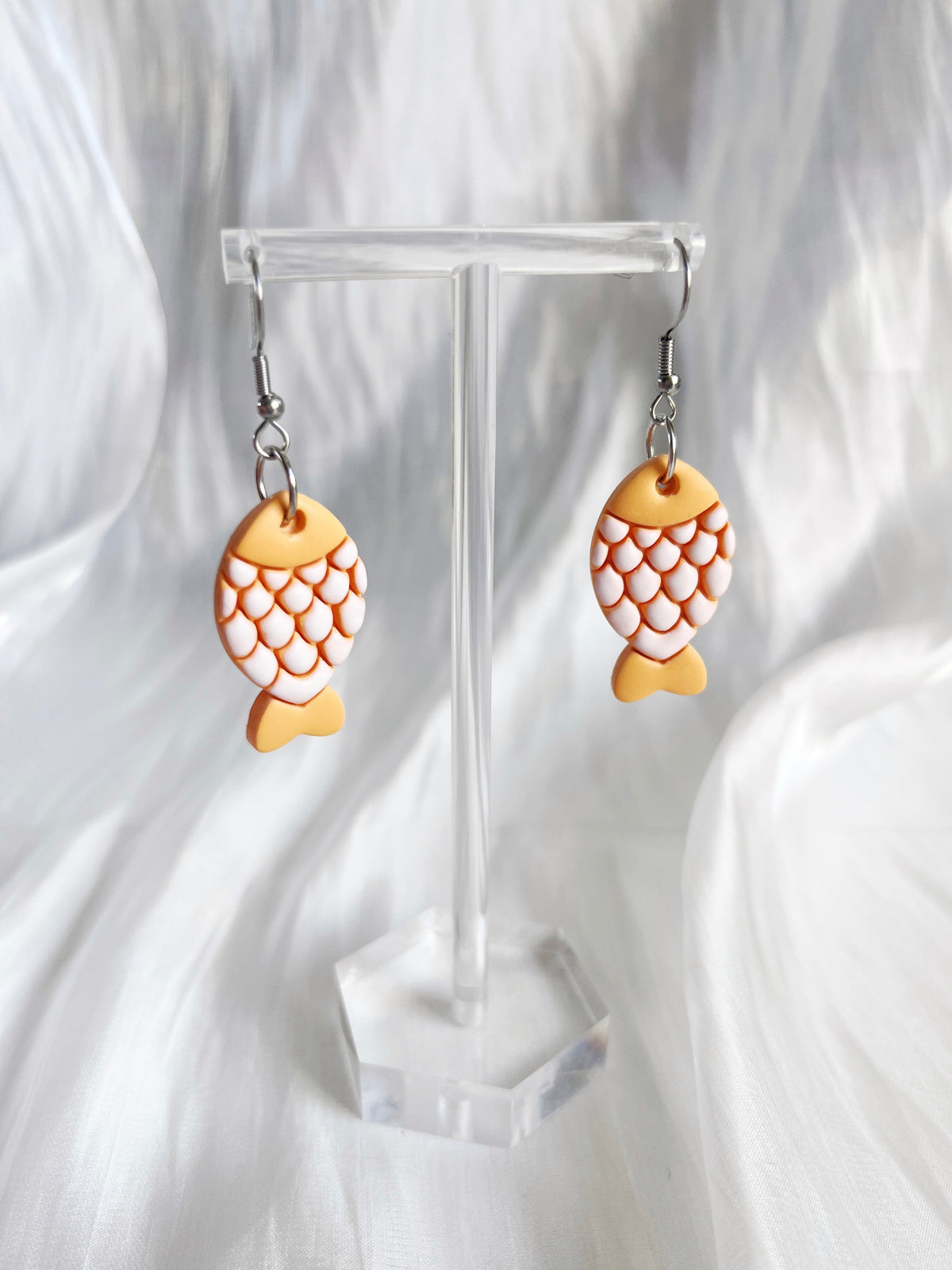 The Fishy Polymer Clay Earrings by silverstar.nz, featuring orange and white scales, hang on a transparent stand. Their unique fish shape crafted from polymer clay stands out beautifully against a softly draped white fabric backdrop.