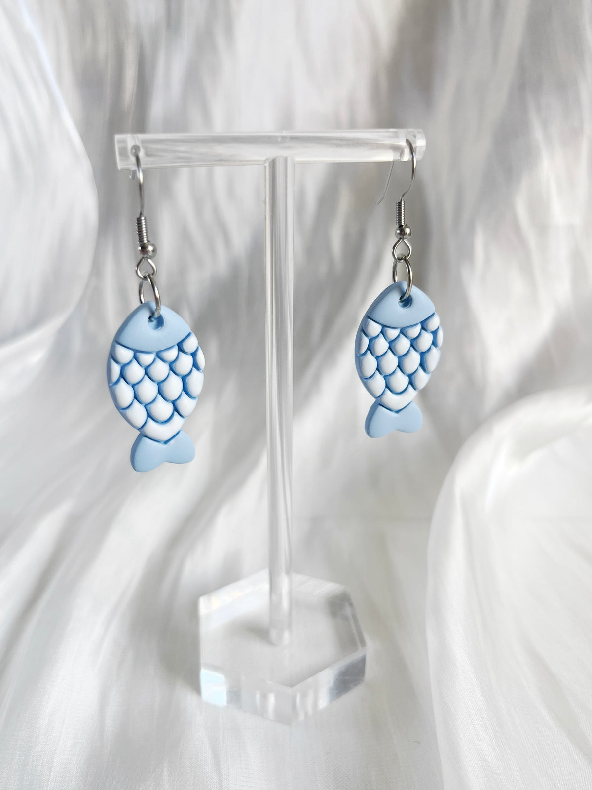 These Fishy Polymer Clay Earrings by silverstar.nz feature blue fish-shaped charms with a scale pattern and hang elegantly from simple hooks, displayed on a clear stand against a wavy white fabric background.