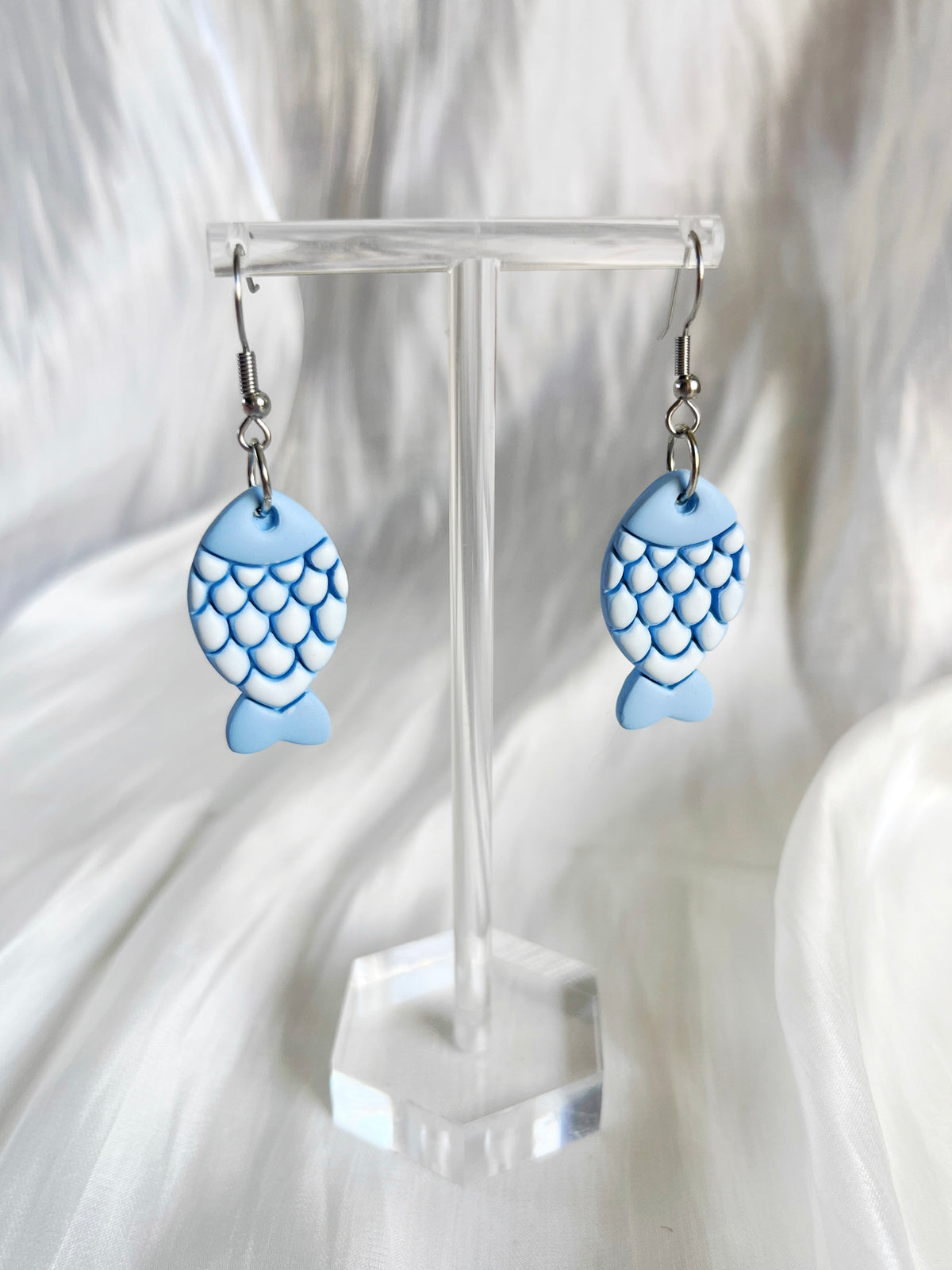 The Fishy Polymer Clay Earrings by silverstar.nz, featuring blue fish-shaped designs with scale patterns, are displayed on a transparent stand against a silky white fabric backdrop.