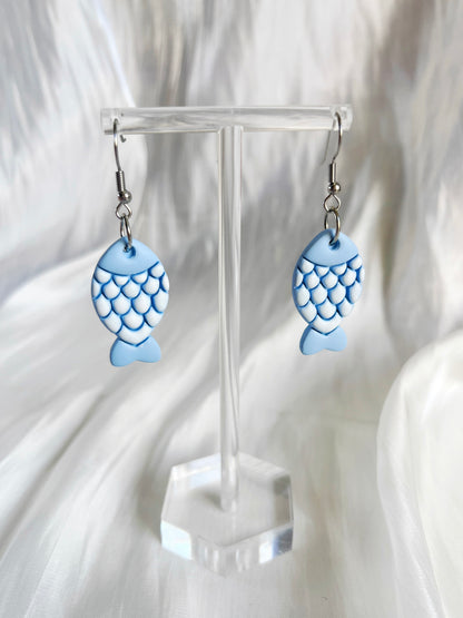 The Fishy Polymer Clay Earrings by silverstar.nz, featuring blue fish-shaped designs with scale patterns, are displayed on a transparent stand against a silky white fabric backdrop.