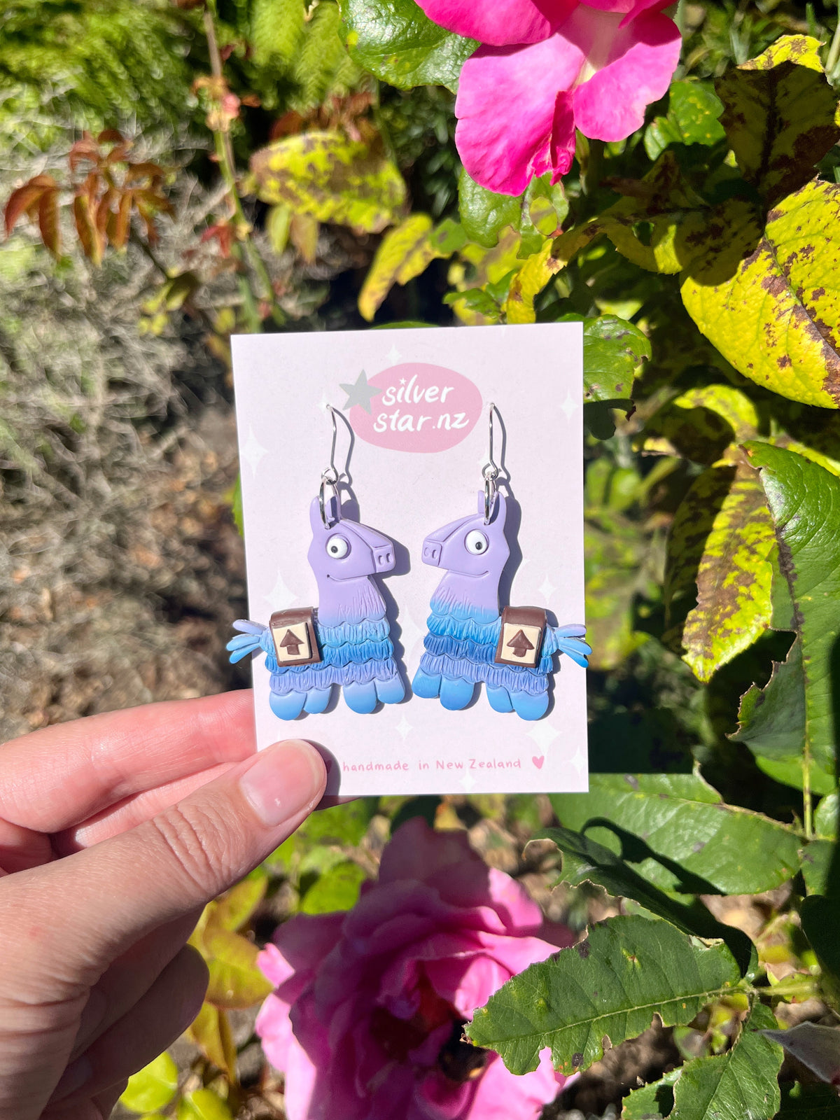 A hand holds a card displaying Llama Polymer Clay Earrings by silverstar.nz in front of green foliage and pink flowers. The card notes they are handmade in New Zealand, highlighting their unique design.