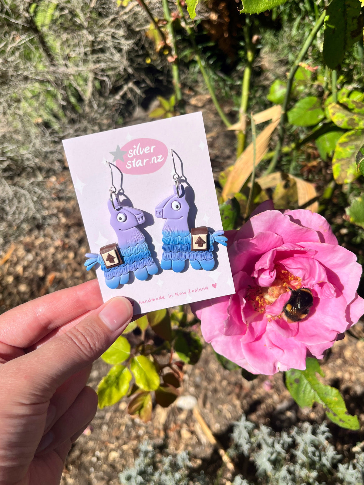 A hand holds a card labeled "silverstar.nz" with handmade Llama Polymer Clay Earrings in front of green foliage and a blooming pink rose, where a bee collects pollen.