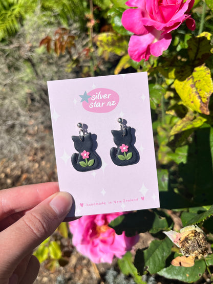 A hand holds a card showing Black Cat Polymer Clay Earrings with pink flowers from silverstar.nz. Pink roses and green leaves are in the sunny background. The card says silverstar.nz and handmade in New Zealand.