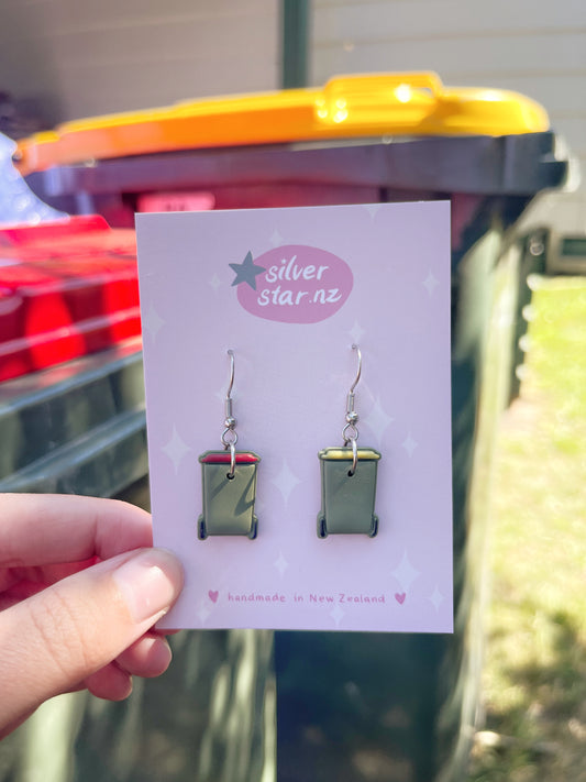 A hand displays silverstar.nz's Rubbish Bin Polymer Clay Earrings—red and yellow bin designs—crafted in New Zealand with durable stainless steel hooks, held before matching outdoor bins.
