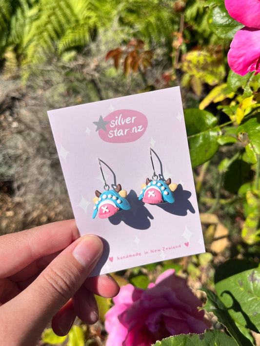 A hand displays a card reading "silverstar.nz" with handmade Chopper Hat Polymer Clay Earrings. Vibrant flowers and green foliage form the backdrop, highlighting these unique earrings crafted in New Zealand.