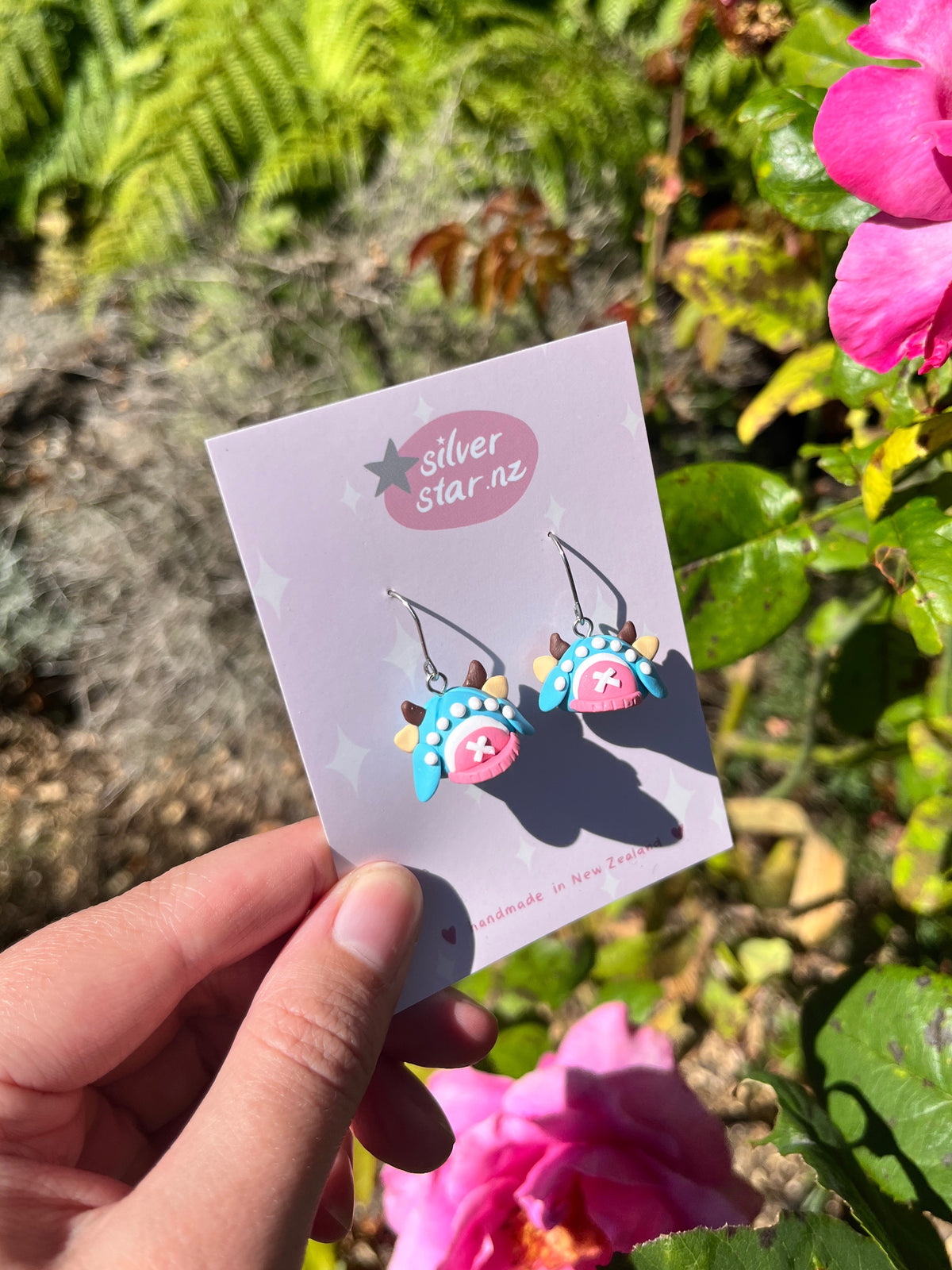 A hand holds a silverstar.nz card displaying colorful, handmade Chopper Hat Polymer Clay Earrings with stainless steel hooks, shown against pink flowers and green foliage in bright sunlight.