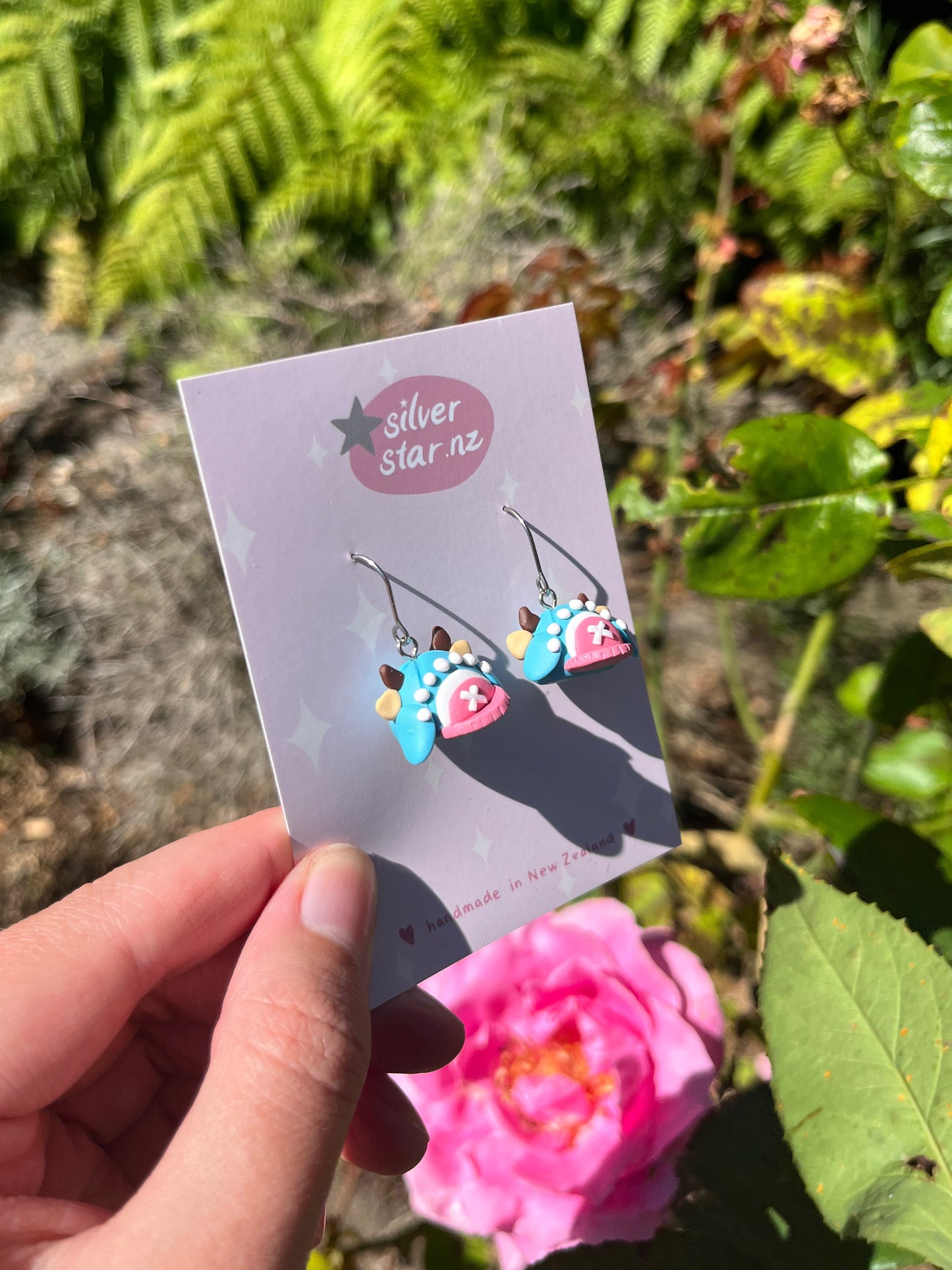 A hand holds a card from silverstar.nz featuring Chopper Hat Polymer Clay Earrings in light blue with pink and white details. Sunlight highlights a pink rose and green foliage in the background.