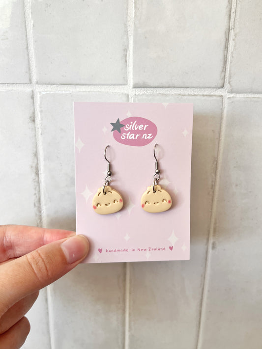 A hand displays Bao Bun Polymer Clay Earrings with smiling faces on a card. The card reads silverstar.nz and handmade in New Zealand, showcasing stainless steel hooks against a light tile-patterned background.