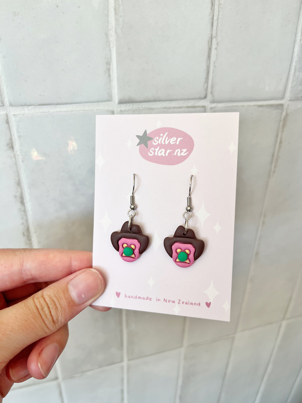 A hand holds a card showing Bubble 'O Will Polymer Clay Earrings by silverstar.nz, shaped like brown cowboy hats with pink brims and turquoise-green flowers, crafted with stainless steel hooks and handmade in New Zealand.