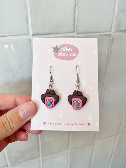 A hand holds a card with Bubble 'O Will Polymer Clay Earrings shaped like iced biscuits, pink borders, sprinkles, and stainless steel hooks. The card shows silverstar.nz and handmade in New Zealand in soft pastel colors.