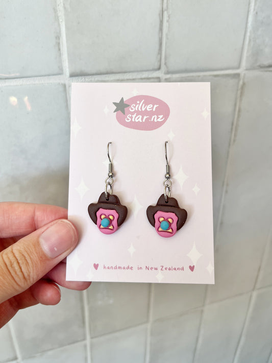 A hand holds a card with Bubble 'O Will Polymer Clay Earrings shaped like iced biscuits, pink borders, sprinkles, and stainless steel hooks. The card shows silverstar.nz and handmade in New Zealand in soft pastel colors.