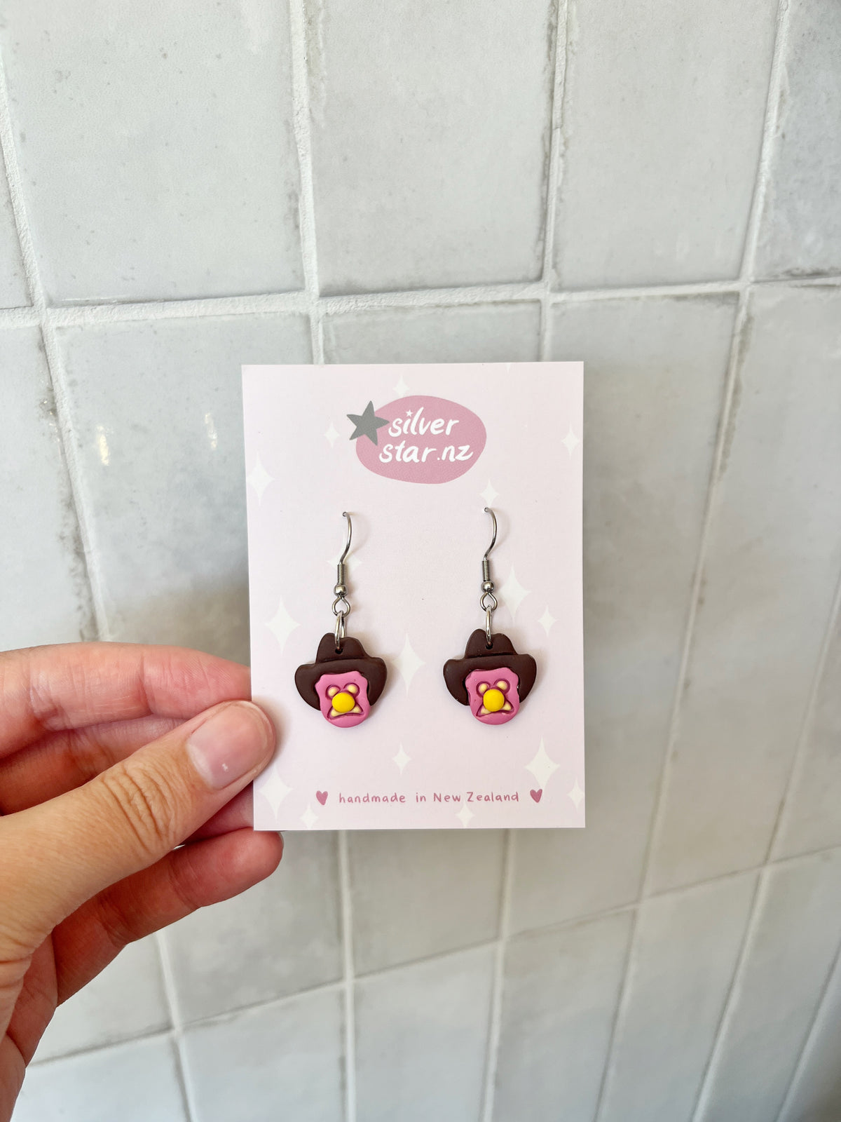 A hand holds a card with Bubble 'O Will Polymer Clay Earrings by silverstar.nz, featuring handmade brown bears with pink and yellow details. The card reads "silverstar.nz" and "handmade in New Zealand," shown against a tiled background.