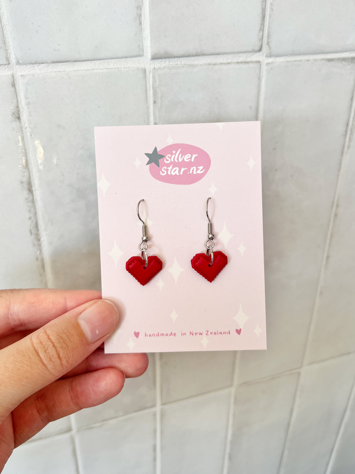 A hand holds a card labeled silverstar.nz with Pixel Heart Polymer Clay Earrings—red heart-shaped handmade pieces on stainless steel hooks, crafted in New Zealand, shown against a tiled background.